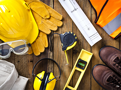 Personal protective workwear and blueprint with some measuring instruments shot directly from above on rustic wood background. The protective workwear includes hard hat, gloves, earmuff, goggles, steel toe shoes, and safety vest. The composition also includes a tape measure and bubble level and a construction blueprint, all items used by construction worker or engineer. Predominant colors: yellow and brown.